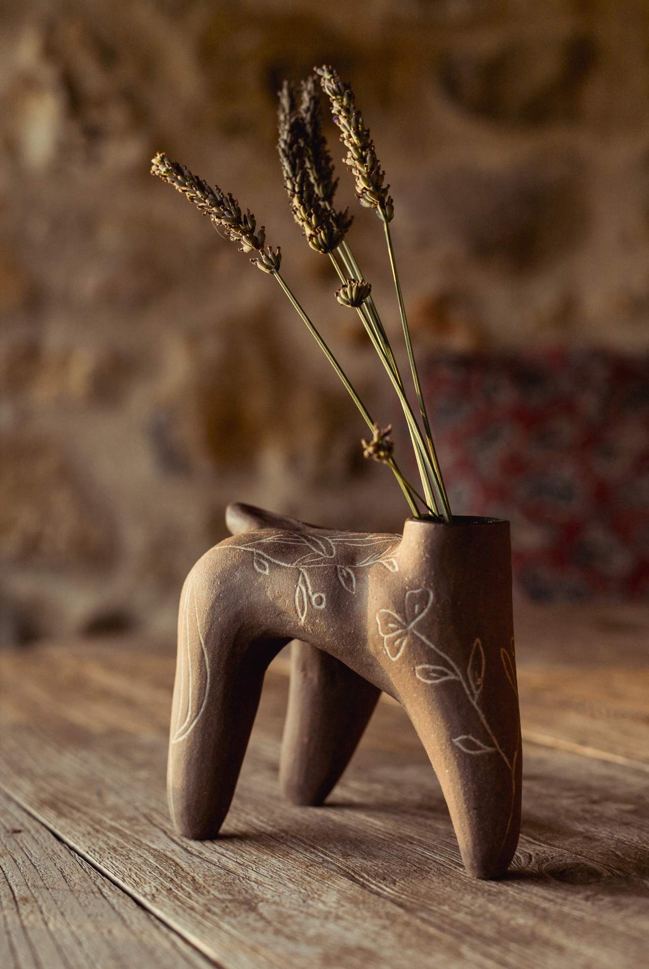 Pièce brune à quatre pieds, feuillages incisés, tenant des épis de graminées séchées, sur une table en bois devant un mur de pierre.