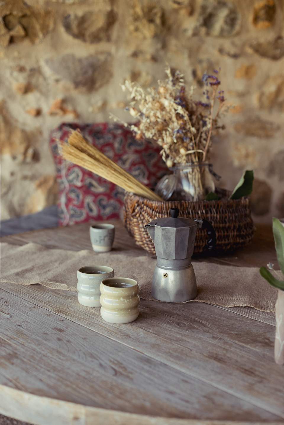 Table ronde en bois : tasses, cafetière italienne, panier de fleurs séchées, devant un mur de pierre.