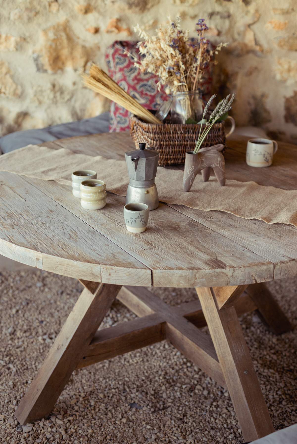 Table ronde en bois vue de haut, tasses et cafetière sur une toile de lin, sol de gravier.